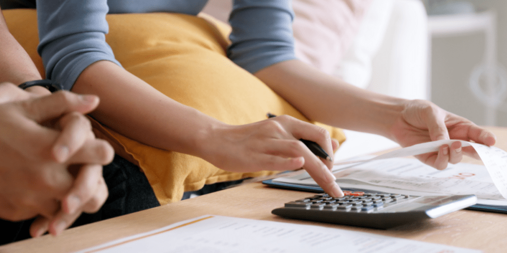 Two people in a home setting, sitting at a coffee table looking at utility bills, with one using a calculator and holding a pen and a receipt.