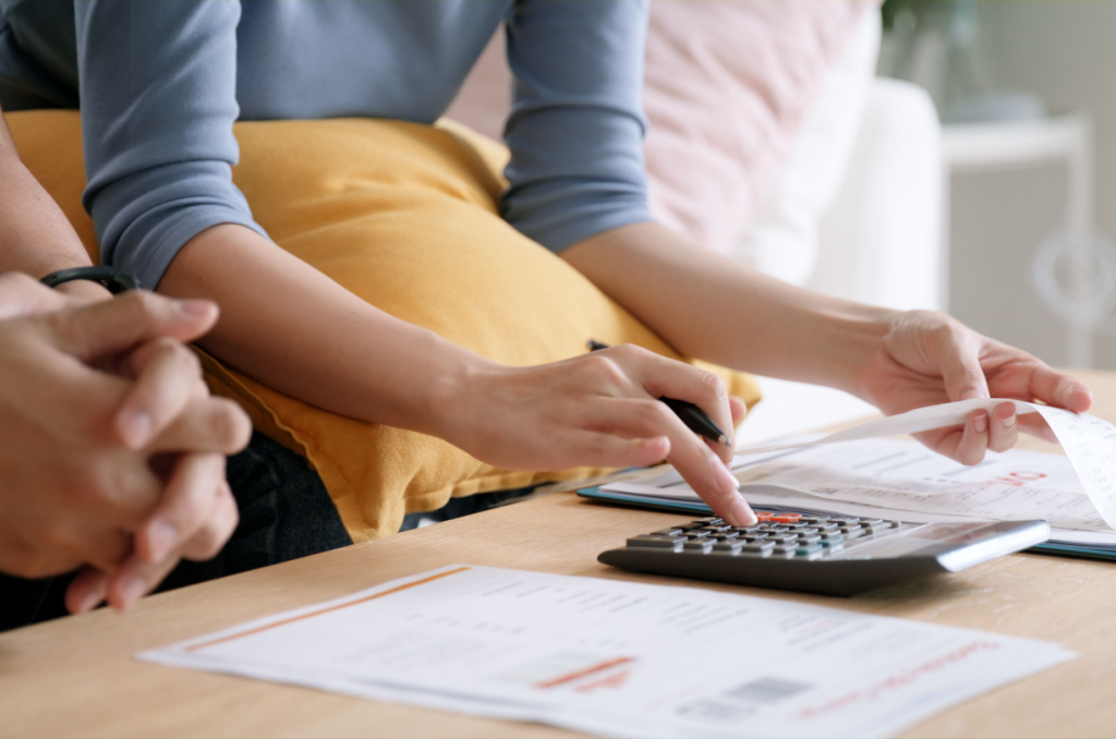 Two people in a home setting, sitting at a coffee table looking at utility bills, with one using a calculator and holding a pen and a receipt.