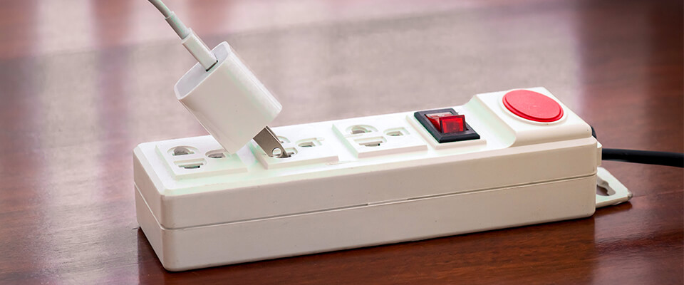 White power strip with multiple outlets on a dark wooden surface, featuring a red power button and a black=red switch, with a white charger being plugged in or unplugged.