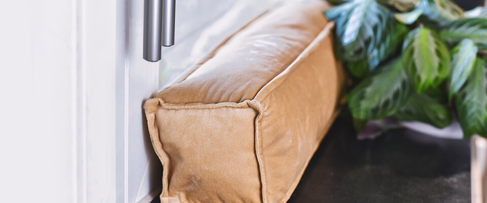 Light brown draft excluder placed on a windowsill, next to a green plant, preventing cold air from entering and saving energy.