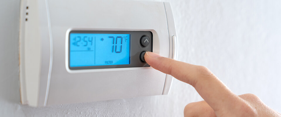 Person adjusting a digital thermostat mounted on a white wall, displaying a temperature of 70°F with a blue-lit screen.