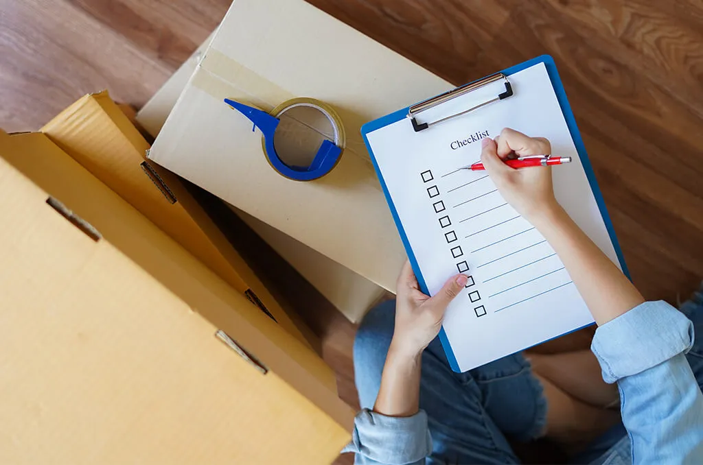 A person sitting on a wooden floor with a checklist on a clipboard, writing with a red pen, surrounded by cardboard boxes and packing tape.