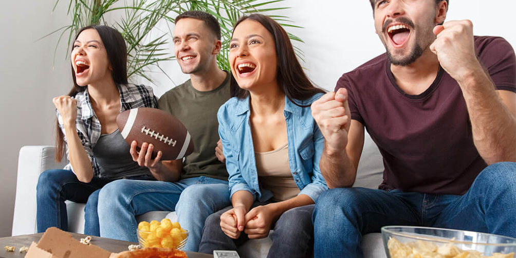 Group of four friends sitting on the sofa, looking excited and cheering while watching a football game of tv, in front of a table with snacks.