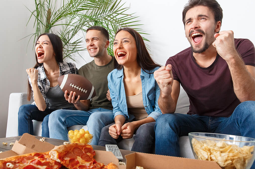 Group of four friends sitting on the sofa, looking excited and cheering while watching a football game of tv, in front of a table with snacks.