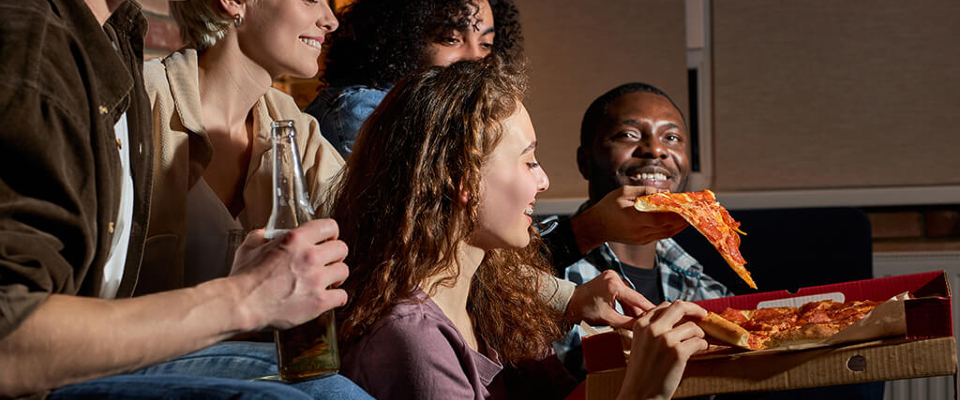 Group of friends smiling and sharing pizza in an indoor setting, with a pizza box open and bottles of beverages in hand.