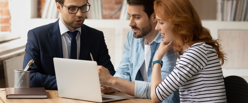 Advisor in a suit discussing renters insurance options with a young couple, in a modern office setting, with a laptop, notebook, and pens on a wooden table.