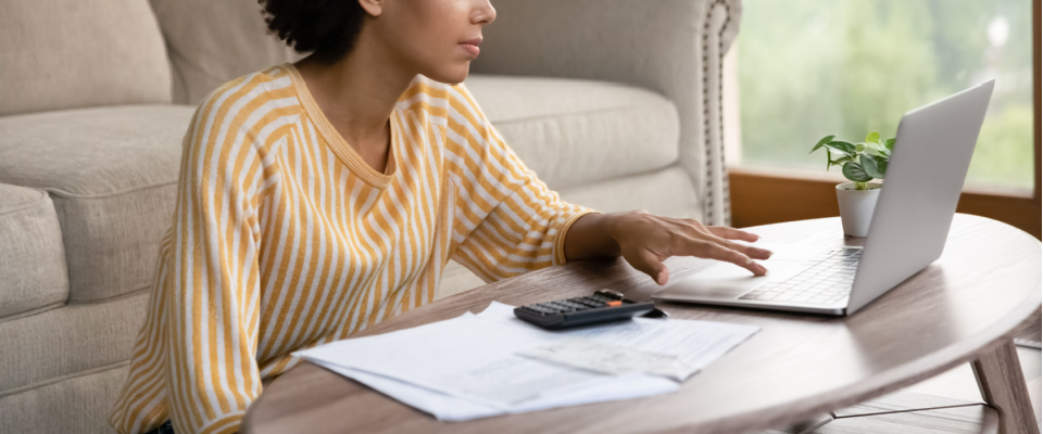 Woman in a striped t-shirt, sitting on the floor in her living room, next to a coffee table with a laptop, calculator, and renters insurance documents.