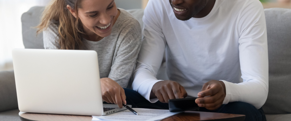 Happy couple sitting on a couch, reviewing renters insurance documents with a laptop and calculator, in a home setting.