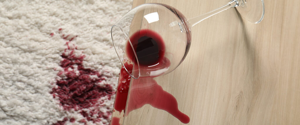 A glass of wine spilling red wine on a white carpet and light wooden floor.