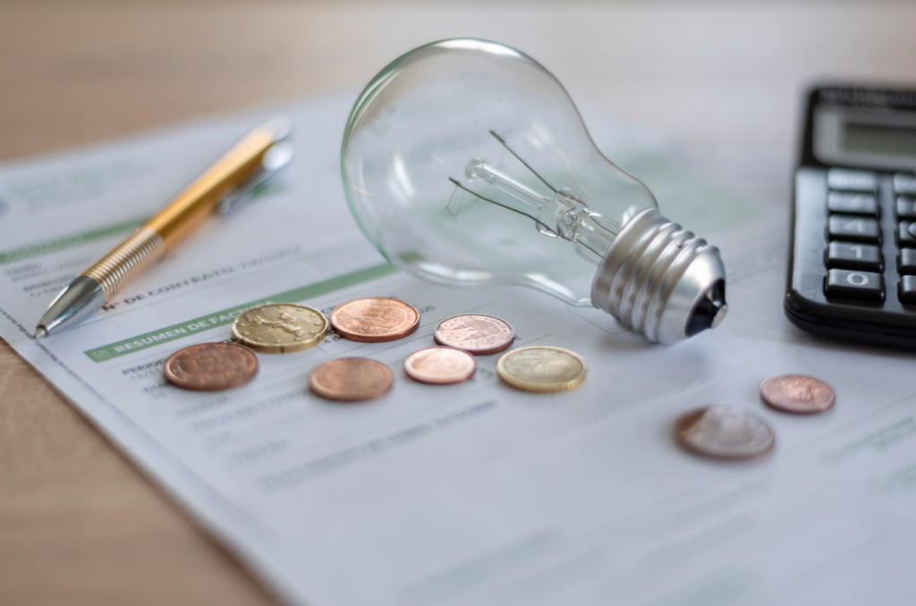 Electricity bills covered by several coins, a light bulb, calculator, and pen on a desk.