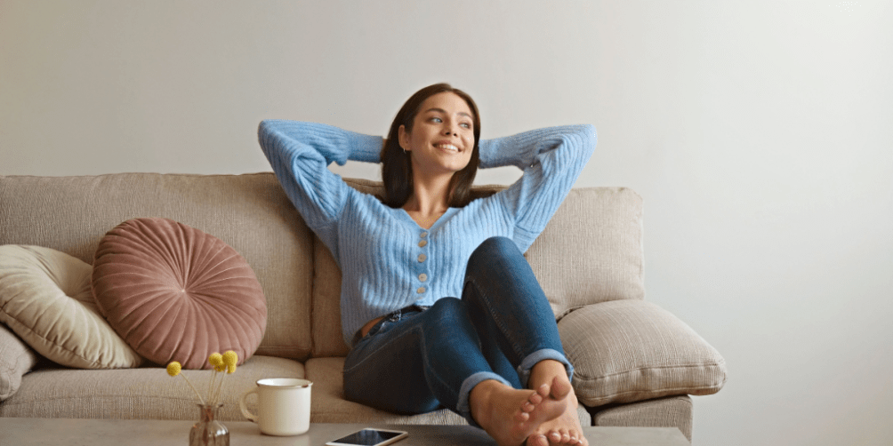 Woman relaxing on beige couch with decorative pillows, wearing a blue cardigan and jeans, feet up on a gray coffee table with a smartphone where she can find renters insurance quotes.