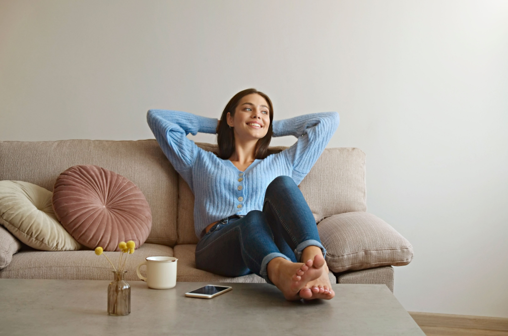 Woman relaxing on beige couch with decorative pillows, wearing a blue cardigan and jeans, feet up on a gray coffee table with a smartphone where she can find renters insurance quotes.