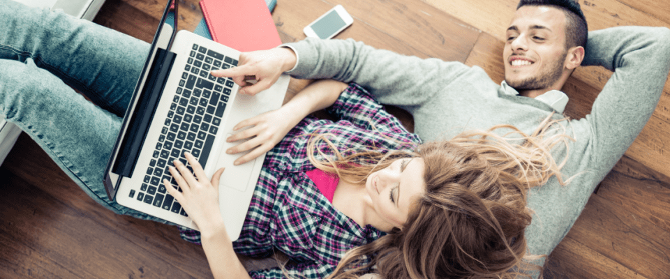 Couple sitting on a wooden floor, looking at a laptop screen searching for renters insurance quotes online, with a smartphone and notebooks next to them.